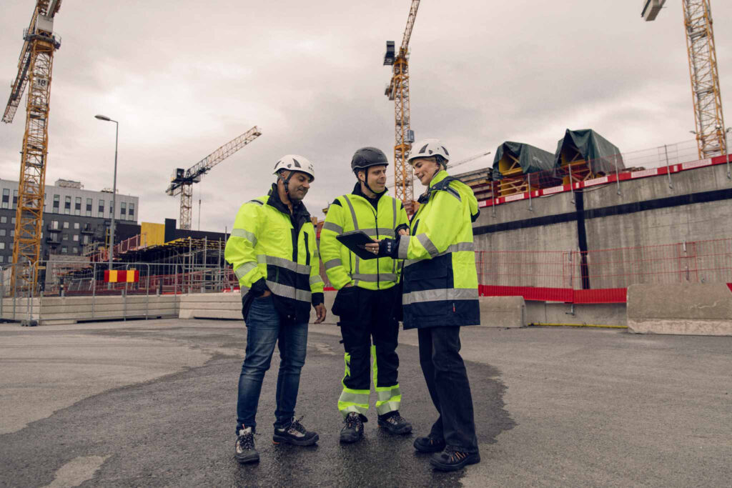 Tre byggarbeidere med verneutstyr diskuterer med en digital enhet på en byggeplass.